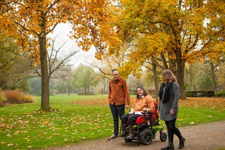 Drei Personen gehen durch einen Park mit bunten Herbstbäumen. Eine Frau sitzt in einem roten Elektrorollstuhl und lacht. Ein Mann und eine Frau gehen neben ihr. Die Blätter auf dem Boden sind gelb und orange. Es ist ein schöner Herbsttag mit frischer Luft.